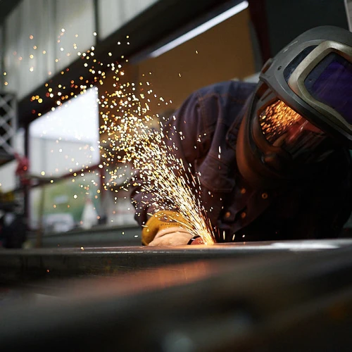 Ein Schweißer arbeitet an einem Metallstück, während Funken fliegen. Der Arbeiter trägt eine Schutzbrille und einen Helm. Hintergrund mit Regalen und Fenstern.