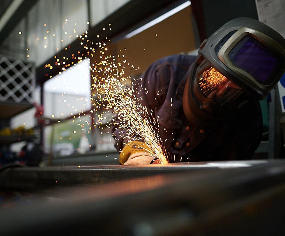 Ein Schweißer arbeitet an einem Metallstück, während Funken fliegen. Der Arbeiter trägt eine Schutzbrille und einen Helm. Hintergrund mit Regalen und Fenstern.