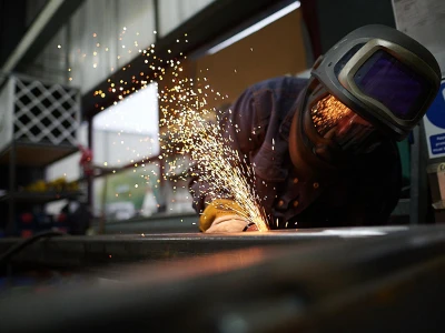 Ein Schweißer arbeitet an einem Metallstück, während Funken fliegen. Der Arbeiter trägt eine Schutzbrille und einen Helm. Hintergrund mit Regalen und Fenstern.