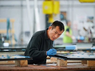 A man wearing a dark sweater and blue gloves works intently at a manufacturing facility, handling large sheets of material on a wooden table. The background features equipment and other workers engaged in similar tasks.