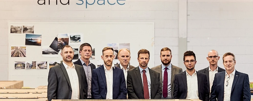 A group of nine professionals stands together in a warehouse, smiling for the camera. They are positioned in front of stacks of wooden crates. A large sign on the wall behind them reads about helping people embrace light, air, and space.