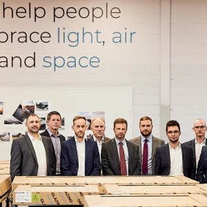 A group of nine professionals stands together in a warehouse, smiling for the camera. They are positioned in front of stacks of wooden crates. A large sign on the wall behind them reads about helping people embrace light, air, and space.