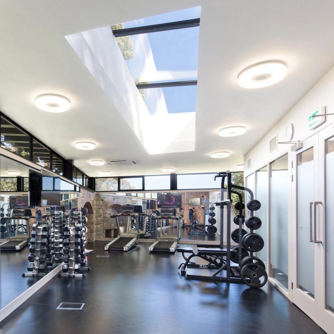 Gimnasio moderno con equipo de entrenamiento, espejos amplios y ventanas grandes que permiten la entrada de luz natural.