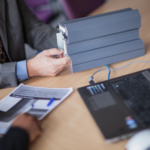 Ein Mann in einem Anzug hält ein technisches Gerät in der Hand, während ein anderer Mann am Tisch eine Dokumentation betrachtet. Auf dem Tisch liegt ein Laptop.