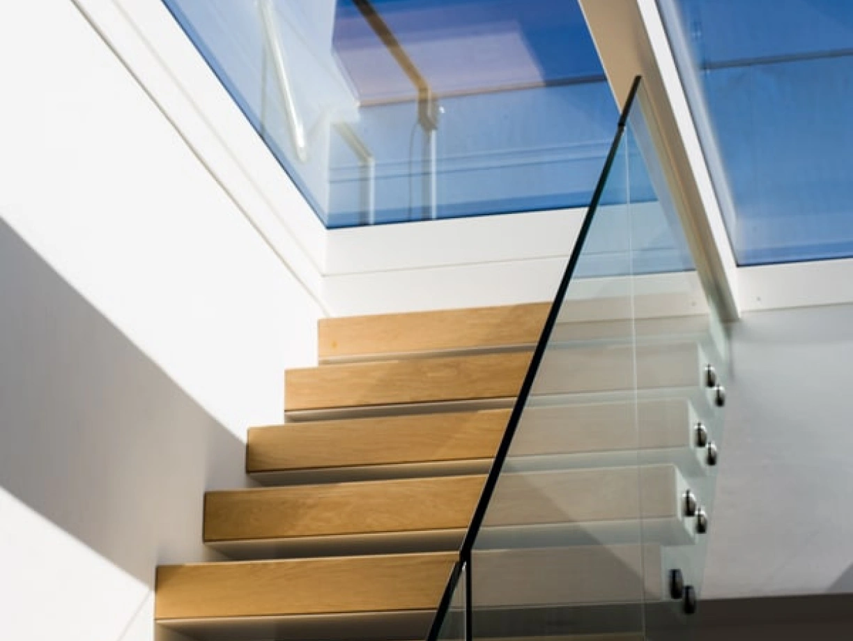 Escalera de madera clara con barandilla de cristal, iluminada por la luz natural que entra por un tragaluz. El fondo muestra un cielo azul despejado.