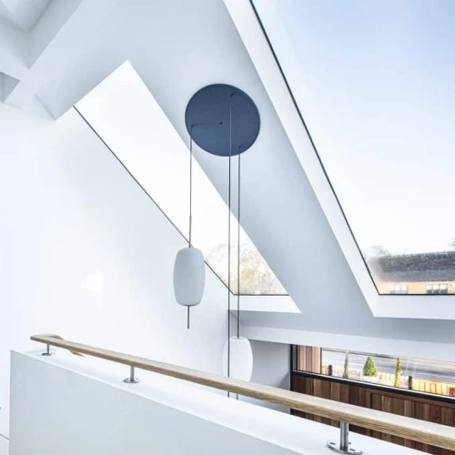 Vestíbulo moderno con techos en ángulo, grandes ventanales y barandilla de madera. Hay un elemento decorativo circular colgante en la pared y una puerta negra que conduce a otra habitación. La luz natural ilumina el espacio.
