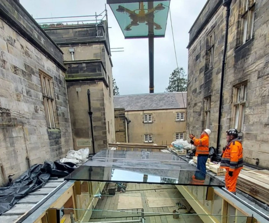 Construction workers wearing safety gear oversee the installation of a glass canopy between two stone buildings, with a large eagle emblem visible in the background.