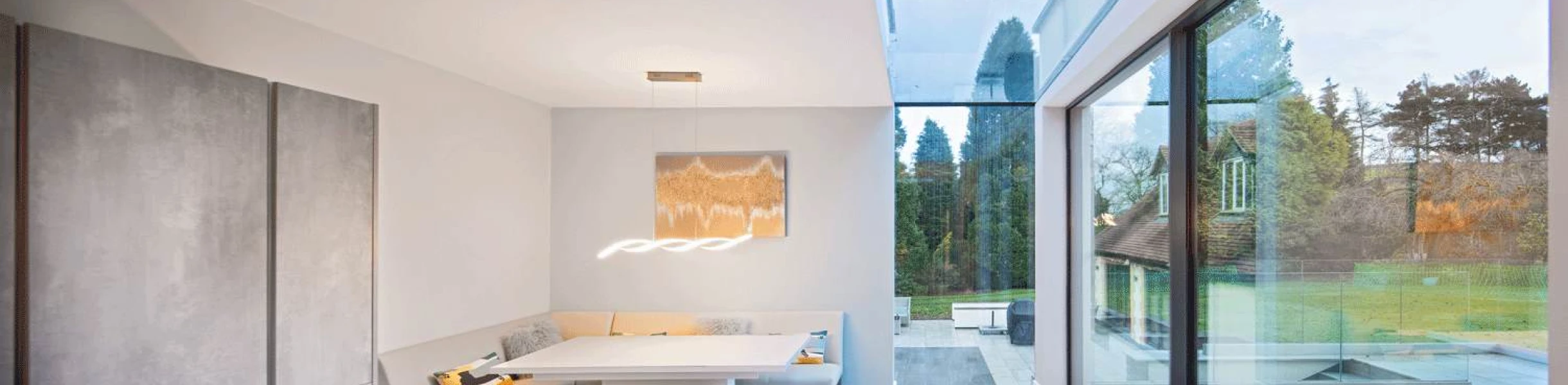 A modern dining area with a minimalist design features a built-in seating nook, a sleek table, and large glass windows showcasing a green outdoor space. Neutral colors dominate the interior, and a stylish light fixture hangs above the table.