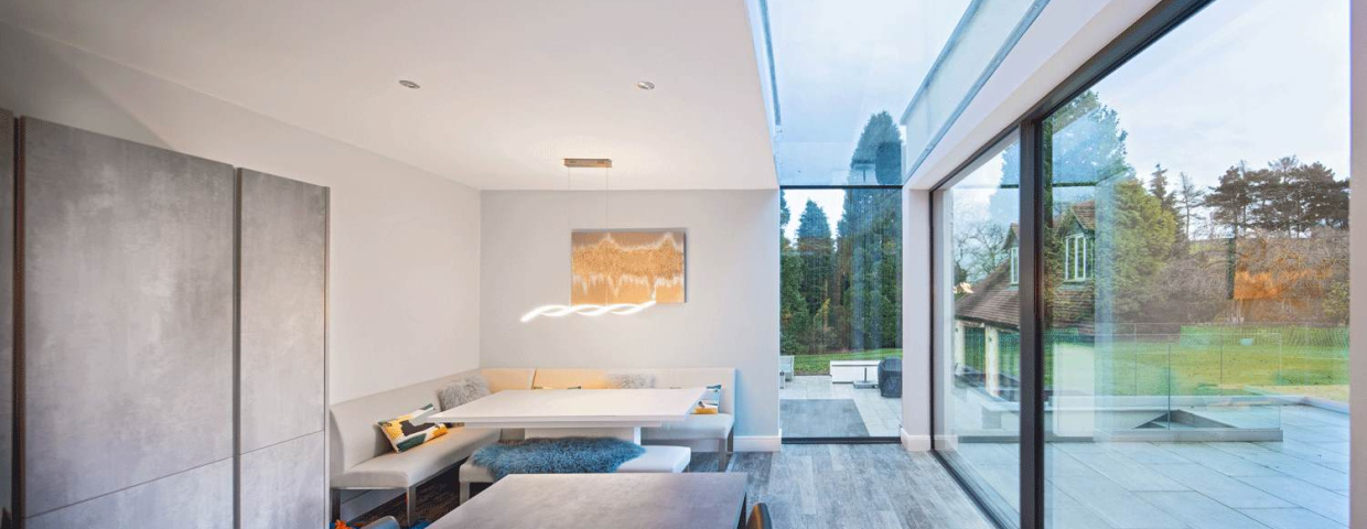 A modern dining area with a minimalist design features a built-in seating nook, a sleek table, and large glass windows showcasing a green outdoor space. Neutral colors dominate the interior, and a stylish light fixture hangs above the table.