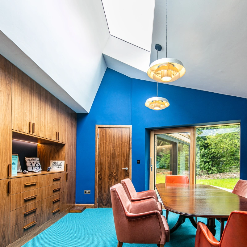 A modern dining room with blue walls, a wooden ceiling, and a large rectangular table surrounded by orange chairs. A large window offers a view of greenery outside. Two stylish light fixtures hang from the ceiling, and a decorative wall clock is visible.