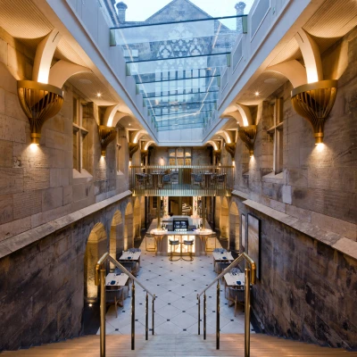 Escalera amplia que desciende a un área con iluminación cálida y paredes de piedra, rodeada de detalles arquitectónicos elegantes. Hay mesas dispuestas en un espacio abierto, con un techo de cristal que permite la entrada de luz natural.