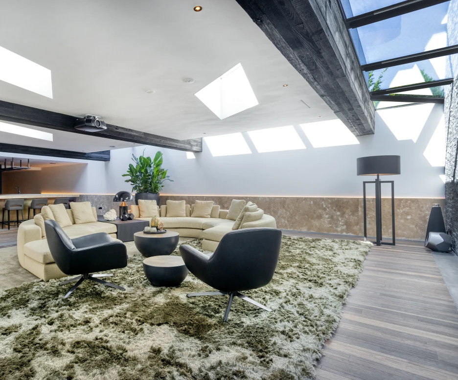 Salon moderne avec un canapé en forme de L, deux fauteuils noirs et un grand tapis moelleux. La pièce est lumineuse grâce aux lucarnes et présente des touches végétales ainsi qu’un bar en arrière-plan.