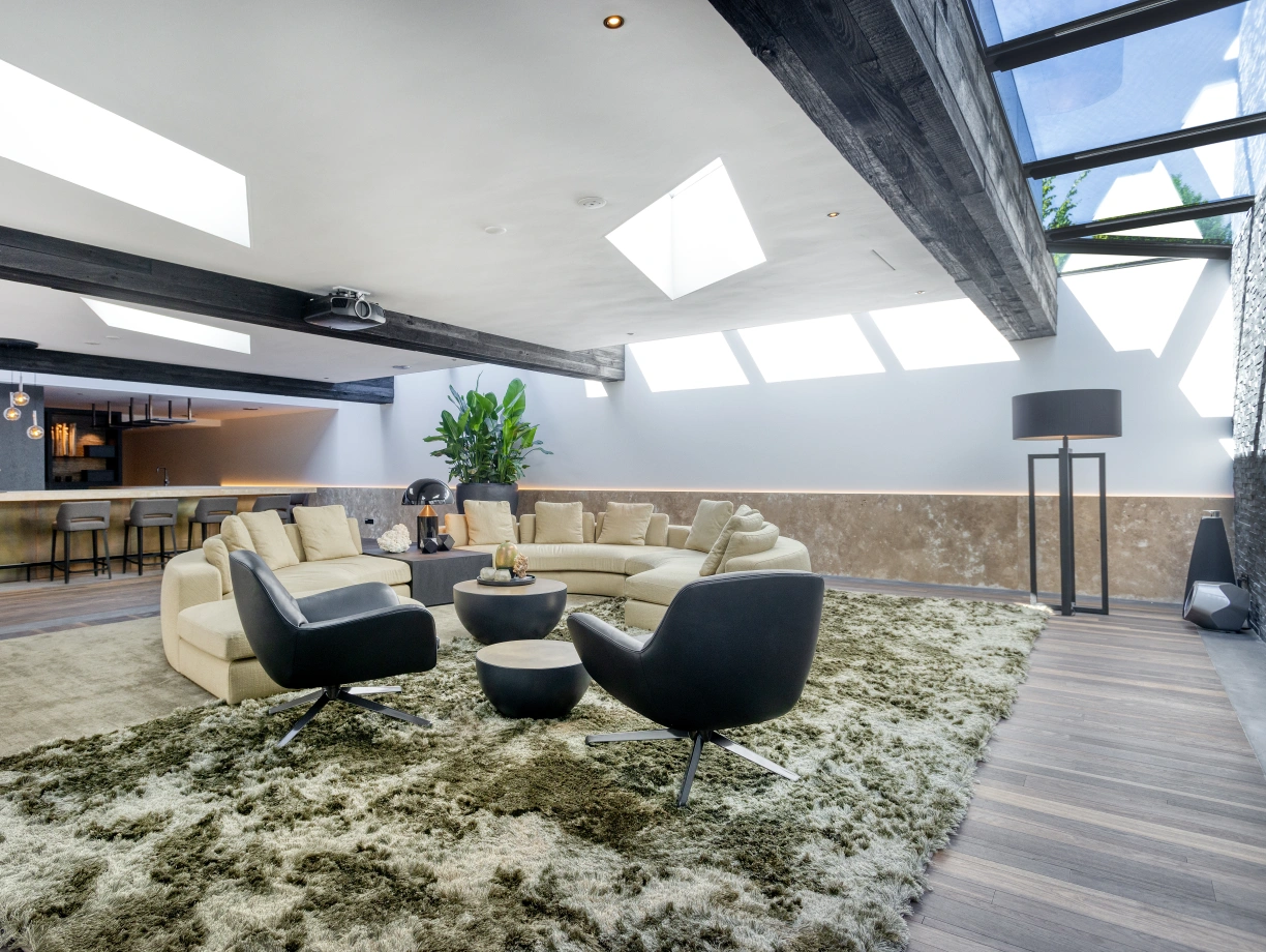 A modern living room featuring a plush, circular sofa, two sleek black chairs, and a round coffee table on a textured rug. Large skylights and a bar area are visible in the background, with greenery adding a touch of nature.