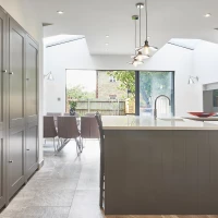 Moderne Küche mit grauen Schränken und einer Kücheninsel. Ein Esstisch mit Stühlen steht im Hintergrund vor großen Fenstern, die auf einen Gartenblick hinausführen. Helle, natürliche Beleuchtung durch Lichtöffnungen im Dach.