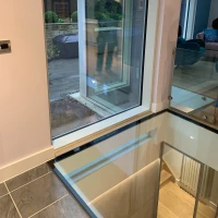 Ventana grande con un marco moderno, mostrando el exterior de una casa y el reflejo de una persona. El área interior cuenta con un piso de baldosas y una escalera de vidrio.