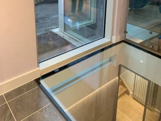 A modern interior with a large window overlooking a garden, featuring a visible reflection of a person standing outside. The floor is made of polished glass, revealing another level below. Soft lighting enhances the contemporary design.