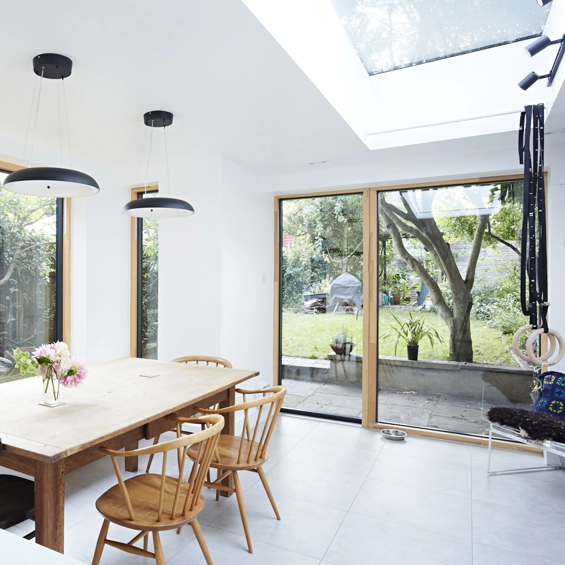 Une salle à manger moderne avec une grande table en bois et des chaises, entourée de fenêtres donnant sur un jardin verdoyant. Deux luminaires noirs suspendus plafonnent l'espace.