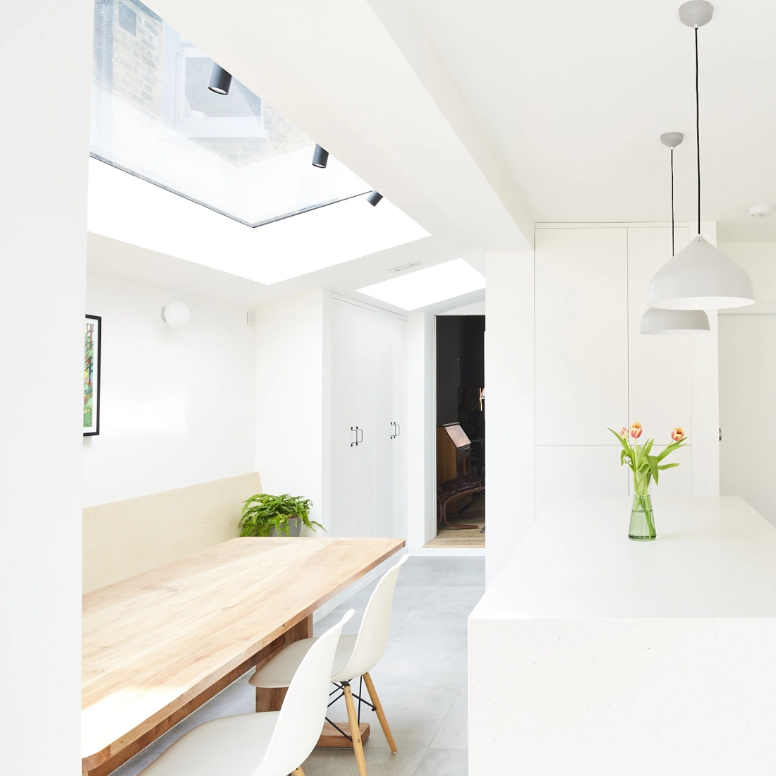 Modern kitchen with white cabinetry, a pink tiled backsplash, and a central island topped with a vase of tulips. A wooden dining table with white chairs is visible, and large skylights provide natural light.