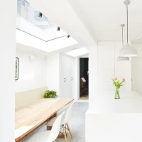 Modern kitchen with white cabinetry, a pink tiled backsplash, and a central island topped with a vase of tulips. A wooden dining table with white chairs is visible, and large skylights provide natural light.