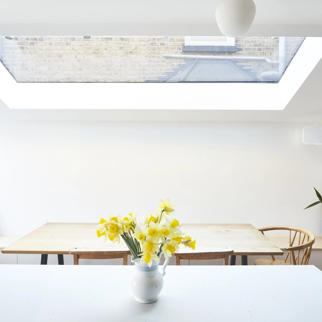Interior de una cocina luminosa con una ventana de techo. En el centro, una mesa de madera rodeada de sillas y un jarrón con flores amarillas. Una planta verde se encuentra en la esquina y hay un cojín decorativo en el fondo.