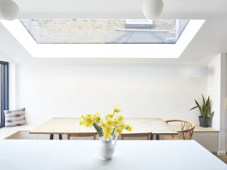 Une cuisine lumineuse avec une grande fenêtre de toit. Une table en bois est placée au centre, entourée de chaises. Un vase blanc contient des jonquilles jaunes sur un comptoir blanc. Une plante verte est visible à droite.
