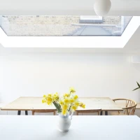 Interior de una cocina luminosa con una ventana de techo. En el centro, una mesa de madera rodeada de sillas y un jarrón con flores amarillas. Una planta verde se encuentra en la esquina y hay un cojín decorativo en el fondo.