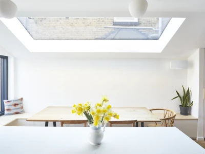 Bright, minimalist kitchen featuring a large skylight, wooden dining table with chairs, and a vase of yellow flowers on the counter. A potted plant adds a touch of greenery.