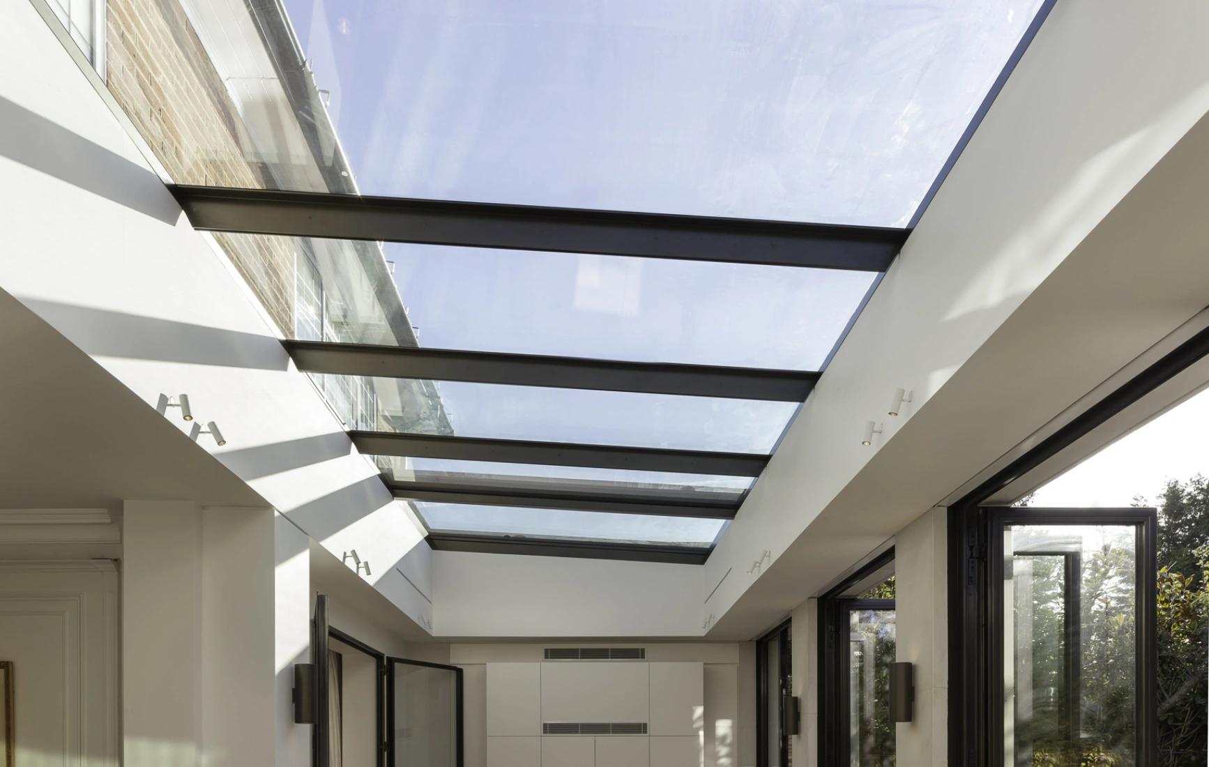 Intérieur lumineux d’une pièce moderne avec un plafond en verre, des portes vitrées ouvertes et un sol en bois. Des rayons de soleil créent des ombres sur le sol, mettant en valeur l’espace