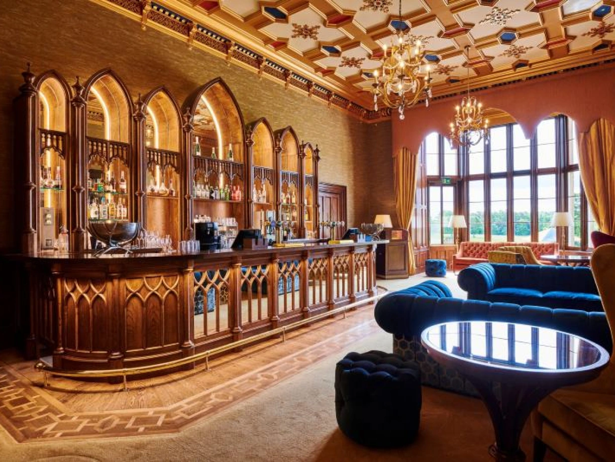 A luxurious bar area featuring an intricately designed wooden bar with shelves of various liquor bottles. The room has ornate ceilings, elegant chandeliers, and plush seating in rich colors. Large windows allow natural light to illuminate the space.