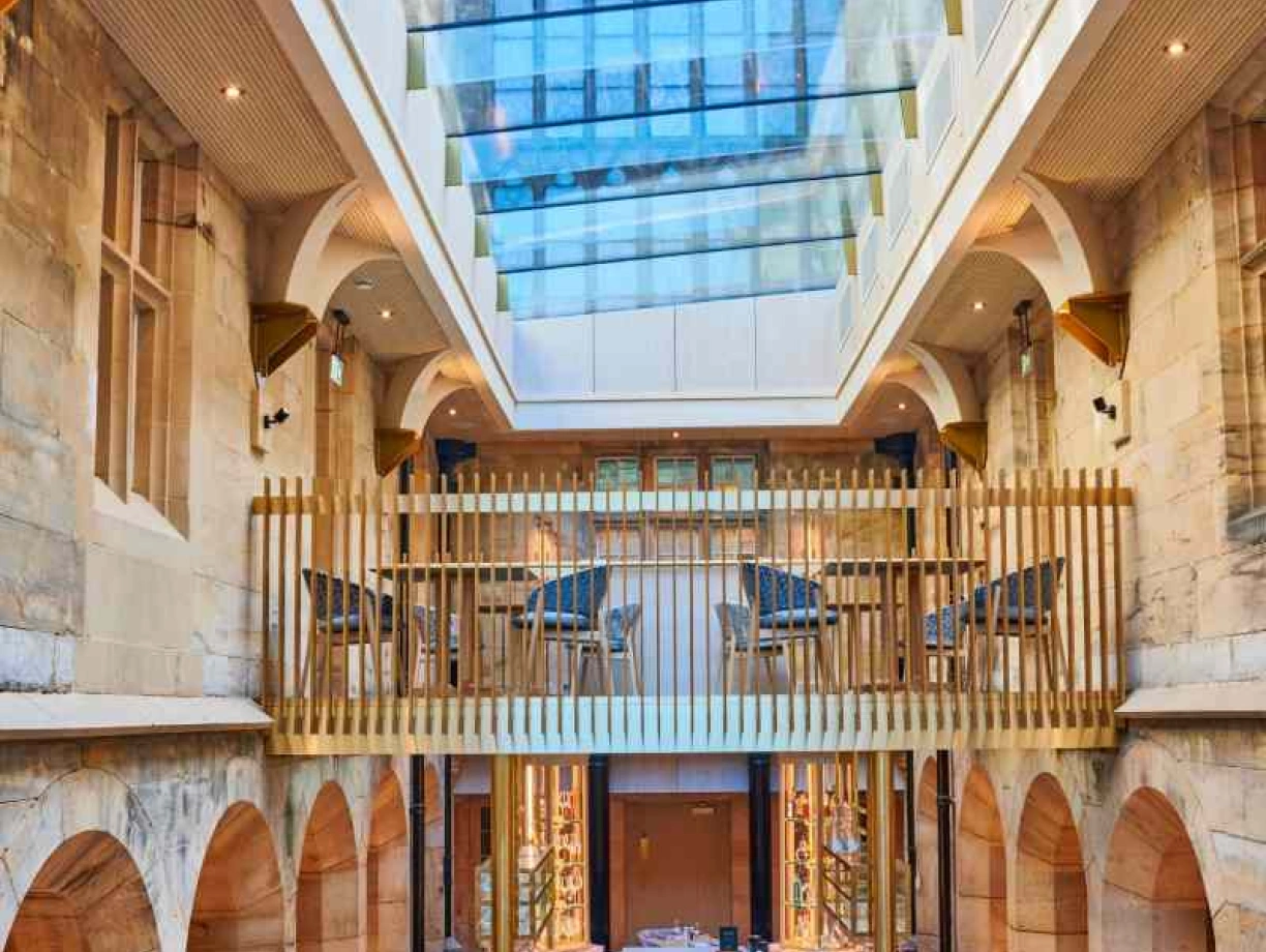 A spacious restaurant featuring a high ceiling with a glass skylight and large arched windows. Wooden beams and a balcony overlook the dining area, which includes neatly set tables and a central bar. The design combines stone walls with contemporary furnishings, creating a bright and inviting atmosphere.
