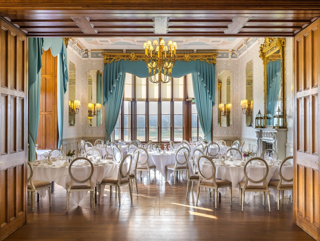 Salón elegante con mesas redondas cubiertas con manteles blancos, sillas de respaldo claro y candelabros adornando el techo. Las ventanas grandes permiten la entrada de luz natural y ofrecen vistas al exterior.