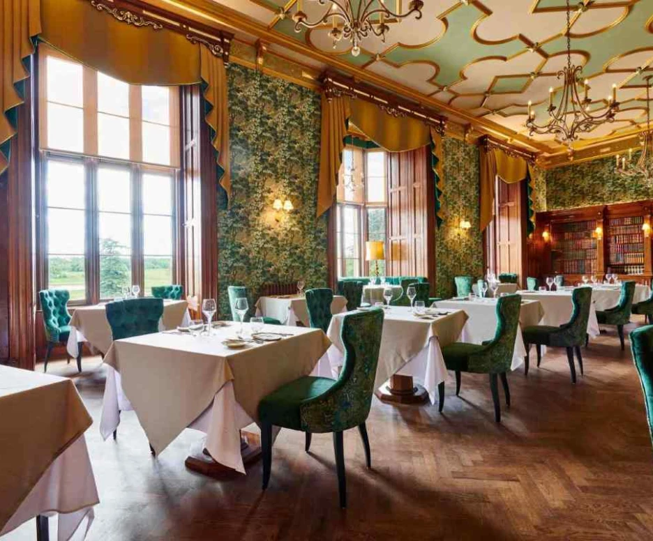 Elegant dining room with green floral wallpaper and ornate chandeliers. Tables are set with white tablecloths and glassware. Large windows provide a scenic view of the outdoors. Bookshelves line one wall, adding to the sophisticated ambiance.