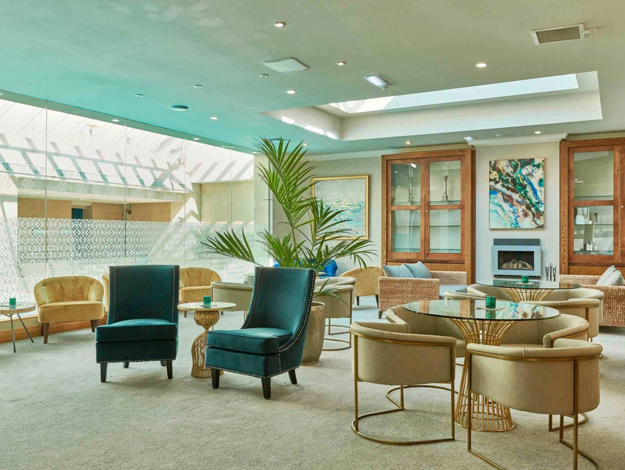 A modern lounge area featuring elegant furniture, including plush chairs and circular tables. Natural light streams in from a skylight, illuminating the neutral-toned carpet and walls adorned with artwork. A decorative plant adds a touch of greenery.