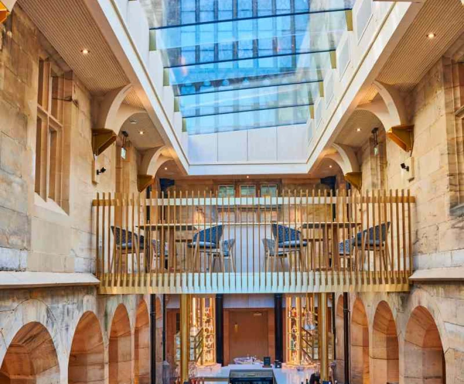 Intérieur d’un restaurant élégant avec un plafond en verre et des arches en pierre. Des tables sont disposées en bas, tandis qu’un bar est visible au centre. Un balcon en bois avec des chaises surplombe l’espace. La lumière naturelle inonde l’ambiance.