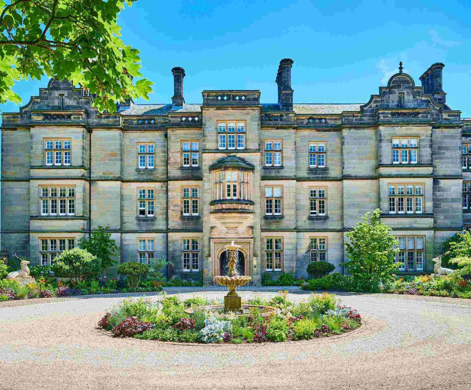 A grand stone building with multiple windows is set against a bright blue sky, surrounded by a circular garden featuring blooming flowers and a central fountain. Lush trees frame the scene, enhancing the picturesque setting.