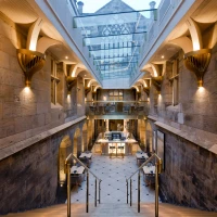 Interiores de un edificio con escaleras de piedra que conducen a un área abierta y elegante, iluminada por luces cálidas y un techo de cristal. Las paredes son de piedra y cuentan con detalles arquitectónicos distintivos.