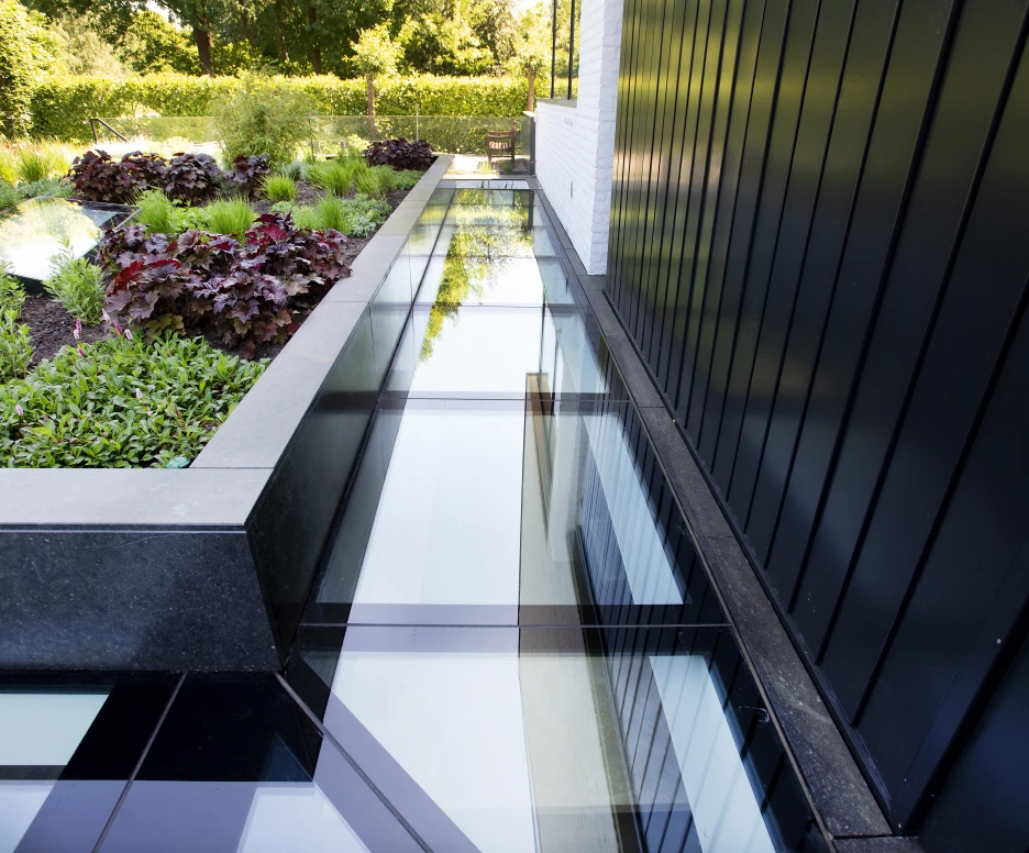 Un chemin en verre transparent borde un jardin verdoyant avec des plantes et des feuillages colorés. Les murs noirs créent un contraste moderne avec la nature environnante.