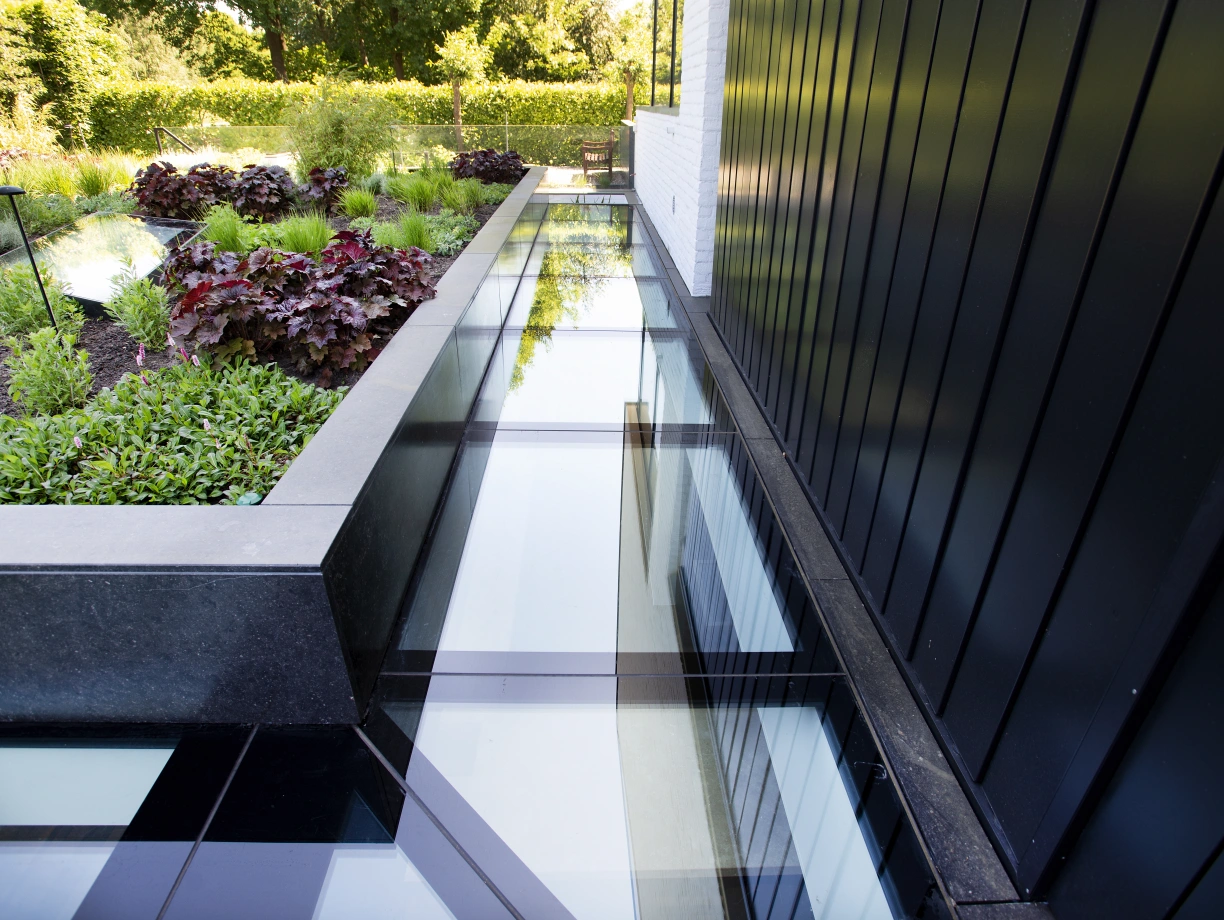 A walkway made of glass panels runs alongside a landscaped area featuring green shrubs and colorful plants. The black exterior of a building is visible in the background.