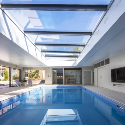 A modern indoor swimming pool features a sleek design with a glass ceiling that allows natural light to flood the space. The pool reflects the surrounding area, which includes sliding glass doors and minimalistic furnishings.