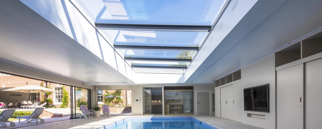Interior elegante con una piscina iluminada por una claraboya. Las paredes son de color claro y hay áreas para relajarse alrededor de la piscina. Se observa un patio exterior a través de las grandes ventanas.