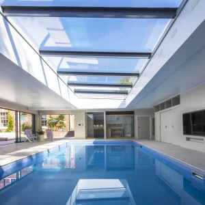 Interior elegante con una piscina iluminada por una claraboya. Las paredes son de color claro y hay áreas para relajarse alrededor de la piscina. Se observa un patio exterior a través de las grandes ventanas.