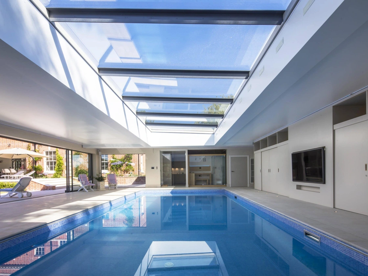 A modern indoor swimming pool features a sleek design with a glass ceiling that allows natural light to flood the space. The pool reflects the surrounding area, which includes sliding glass doors and minimalistic furnishings.