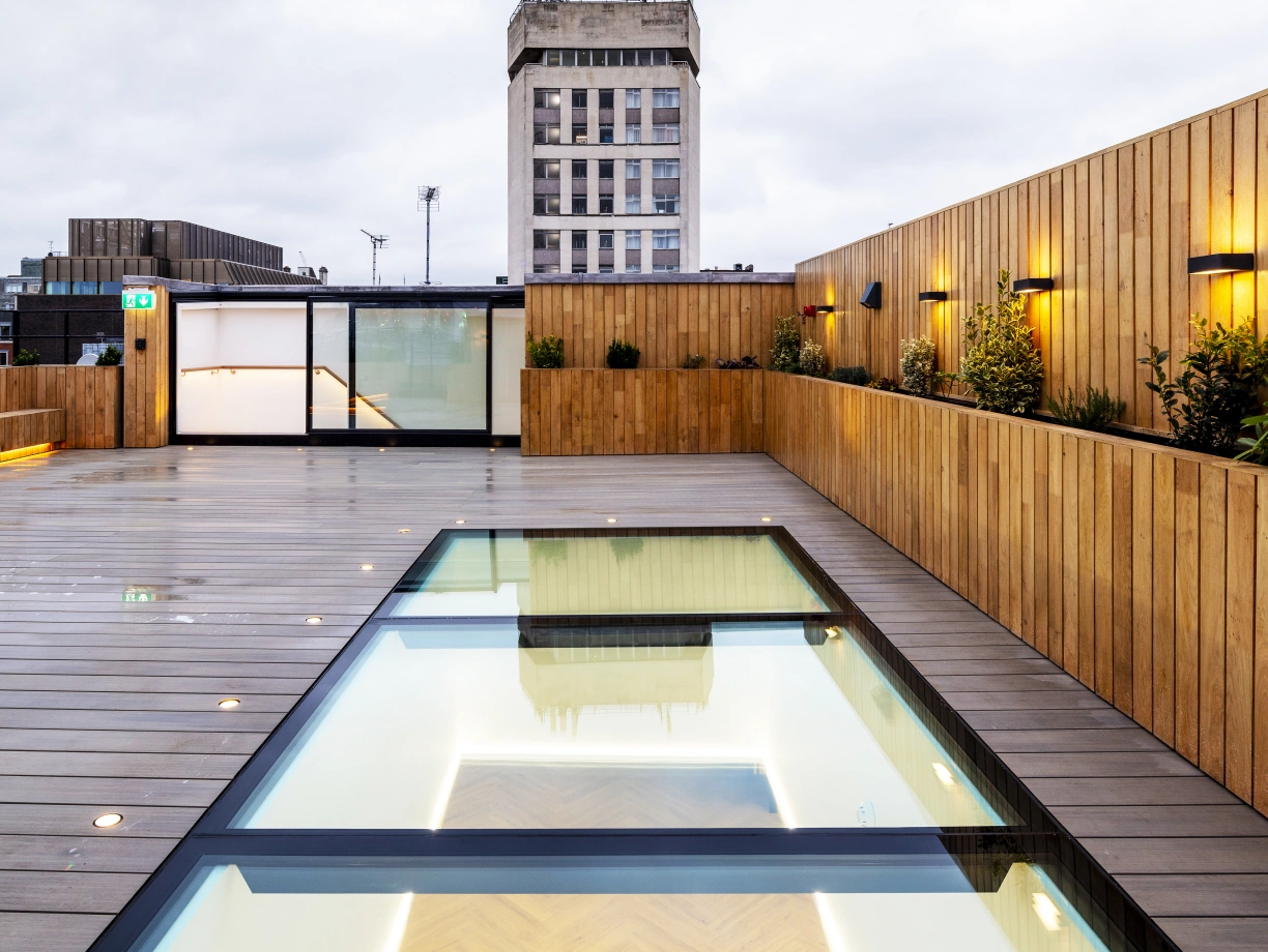 A modern rooftop with a wooden deck and a glass-lined swimming pool reflecting the overcast sky. A tall building stands in the background, adding to the urban atmosphere. Soft lighting accents the space, creating a warm ambiance.