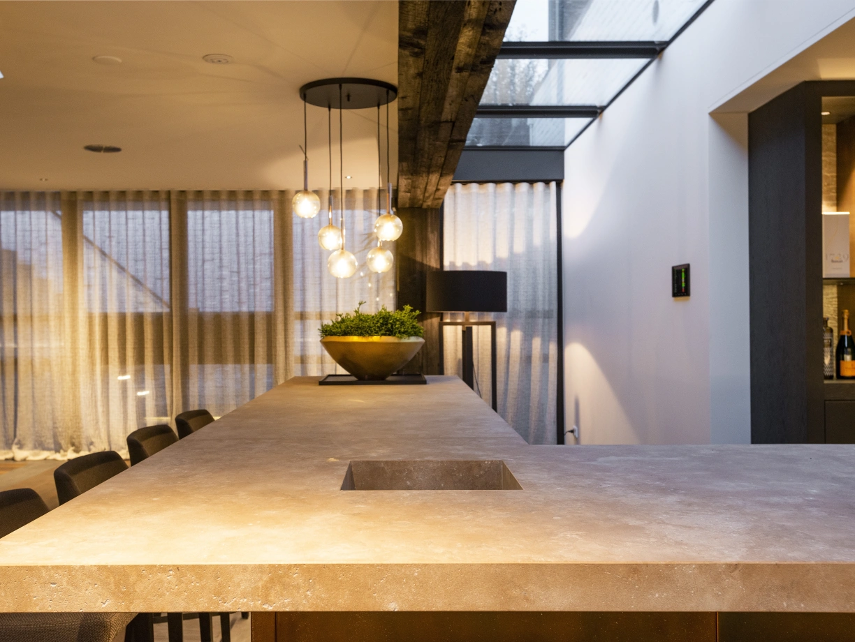 Intérieur d’une cuisine moderne avec un comptoir en béton, un éclairage suspendu élégant et des fenêtres laissant entrer la lumière naturelle. Des rideaux translucides ajoutent une touche de douceur à l’espace.