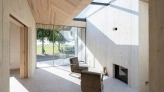 Intérieur d'une maison moderne avec des murs en bois clair et de grandes fenêtres laissant entrer la lumière naturelle. Une chaise en cuir grise est placée face à une cheminée. On aperçoit à l'extérieur une vue sur un espace vert.