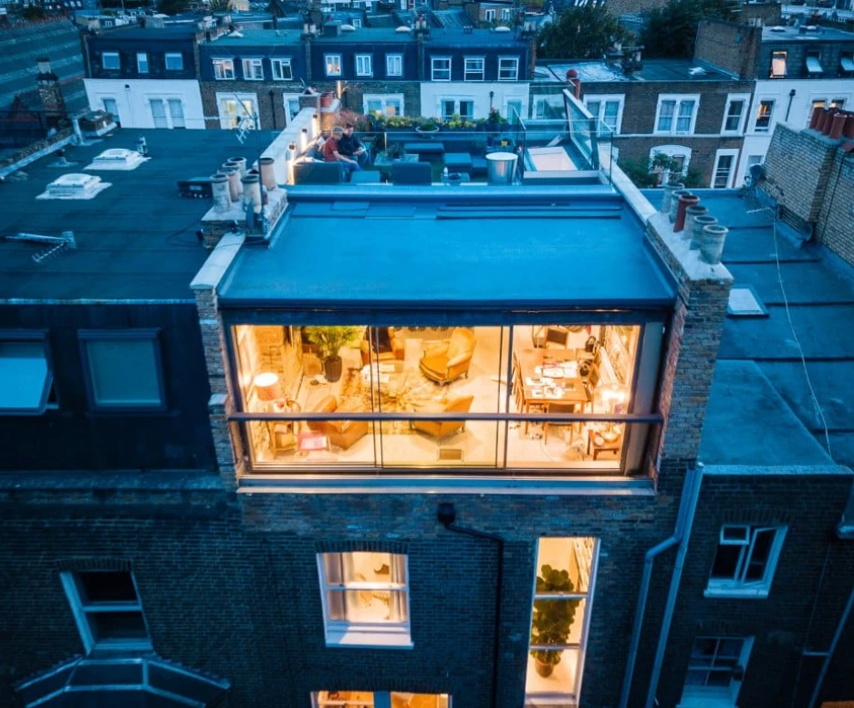 A modern rooftop terrace with a glass-walled lounge area, featuring cozy furniture and decorative plants. Nearby rooftops of city buildings are visible in the background, illuminated by evening lights.