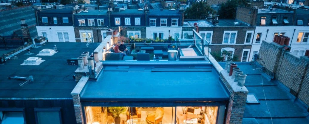 Aerial view of a modern building showcasing a rooftop terrace and large glass windows, revealing a cozy interior with seating and plants. The surrounding rooftops are visible in the background during twilight.