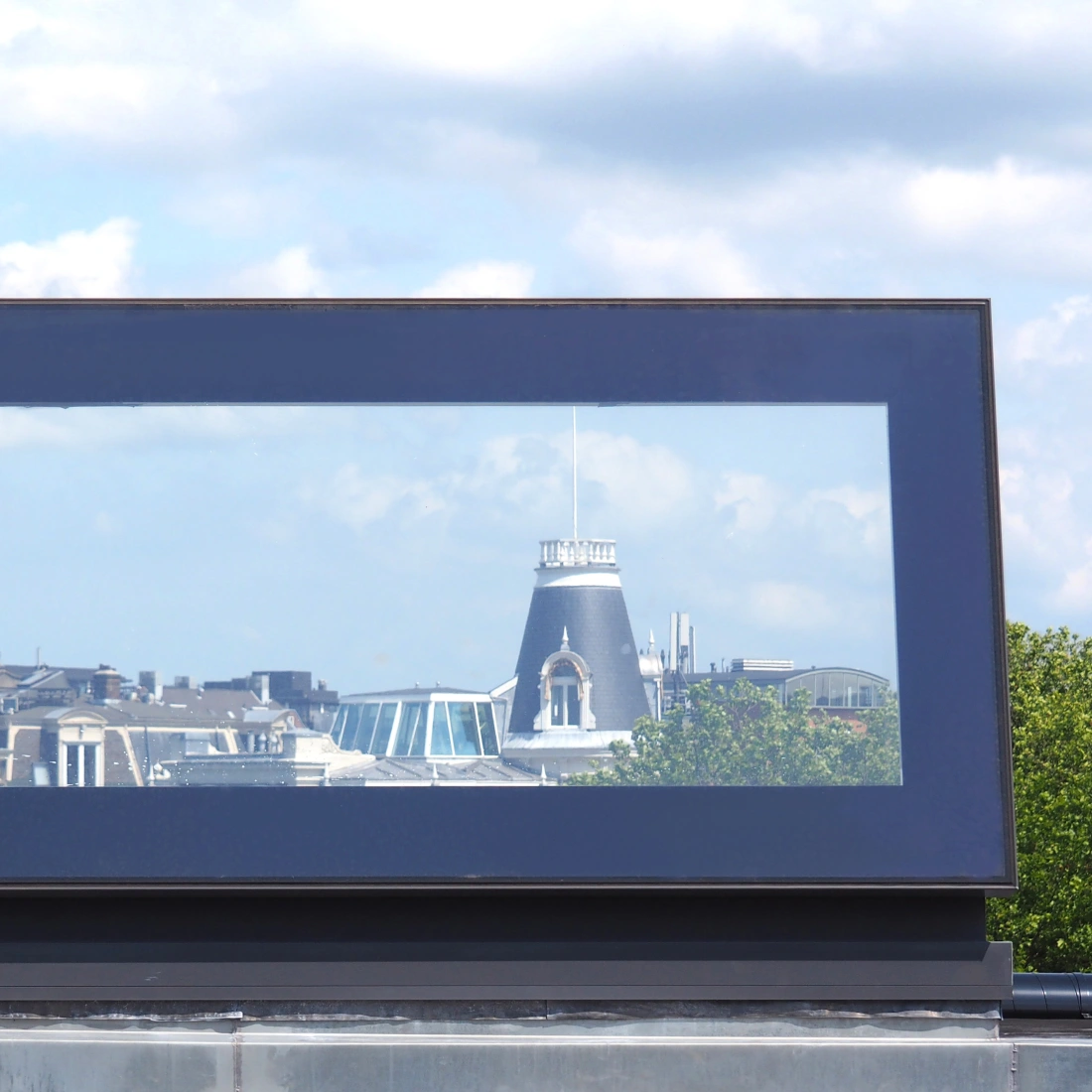 Vue d'un paysage urbain à travers un grand vitrail rectangulaire, montrant des bâtiments historiques sous un ciel partiellement nuageux.