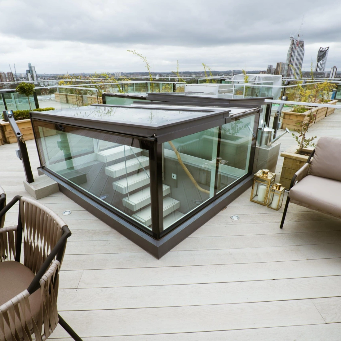 Terraza moderna con barandillas de vidrio, escaleras y muebles de exterior. Se observa una vista urbana con edificios al fondo. El cielo está nublado.