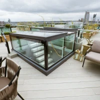A modern rooftop terrace features a glass enclosure and a staircase leading down. Stylish outdoor seating with a table is provided, along with planters and greenery. The skyline and tall buildings are visible in the background under a cloudy sky.